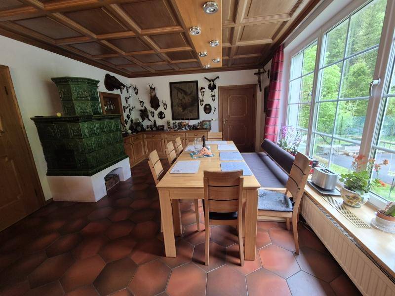 Heller Essbereich mit großem Holztisch, traditionellem Kachelofen und Blick ins Grüne durch große Fenster.