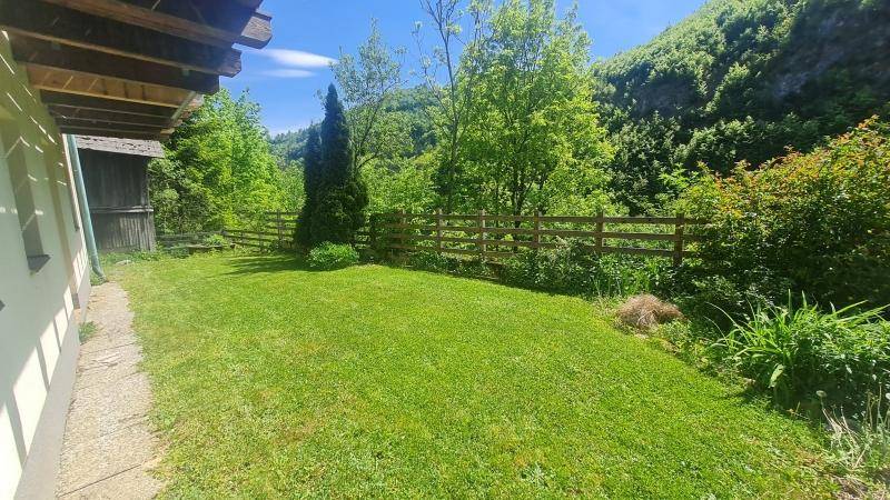 Gepflegter Gartenbereich mit Rasenfläche und Holzzaun, umgeben von Bäumen und grünen Hügeln.