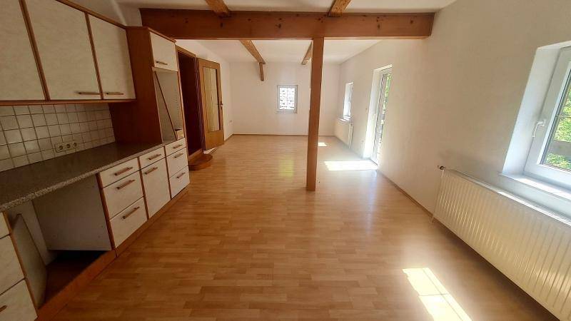 Heller offener Wohnbereich mit sichtbaren Holzbalken, Laminatboden und einer einfachen Küchenzeile.