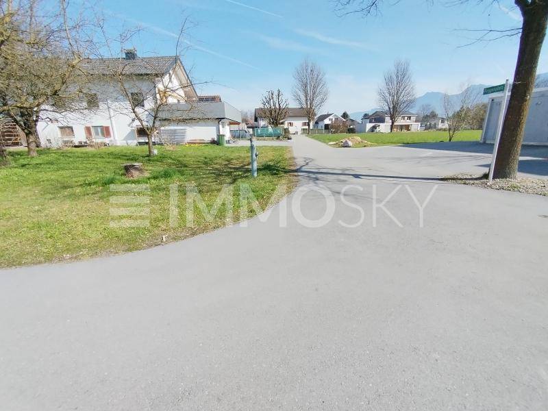 Asphaltierte Zufahrt neben einer Grünfläche mit einem Wohnhaus und Bergen im Hintergrund.