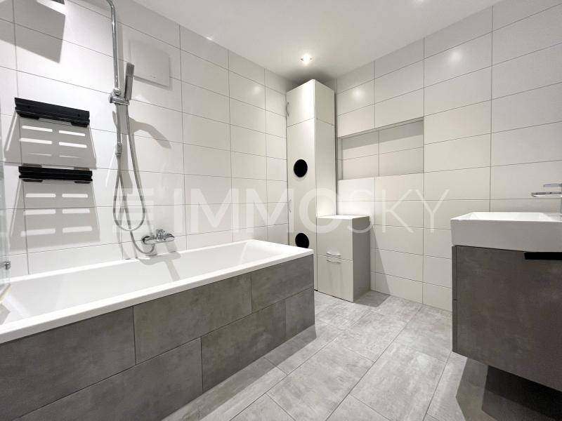 Modernes Badezimmer mit weißen Fliesen, grauen Bodenfliesen, Badewanne und Waschtisch mit Unterschrank.