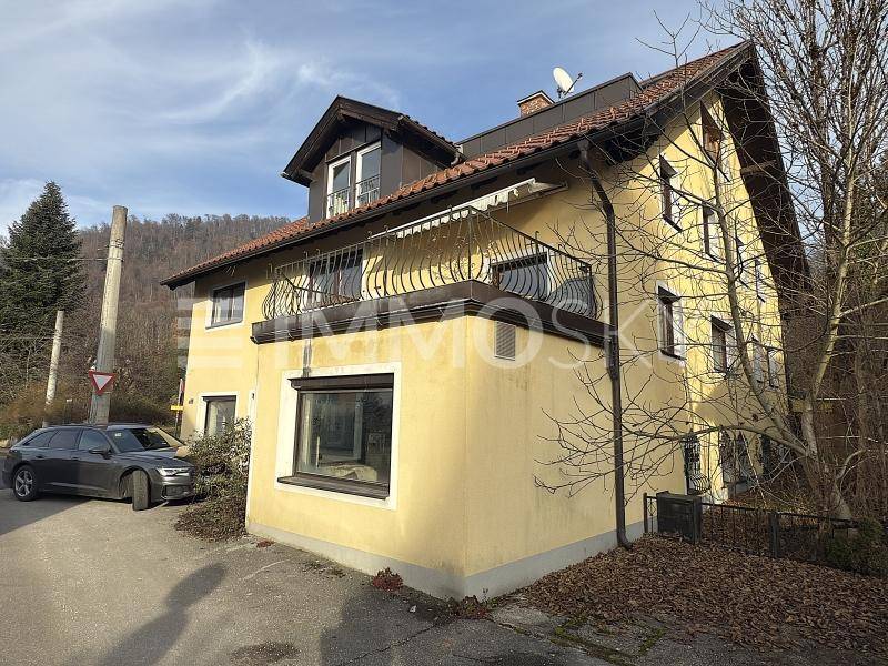 Seitenansicht des gelben Gebäudes mit Balkon, umgeben von Bäumen und einem geparkten Auto.