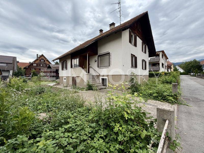 Hausfassade mit Außentreppe zum Eingang und verwildertem Vorgartenbereich.