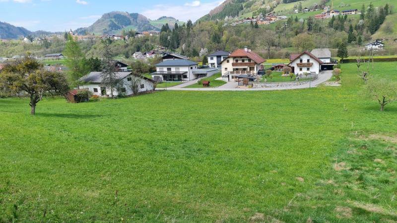 Panoramablick über eine weite grüne Wiese mit modernen Wohnhäusern und Berglandschaft.