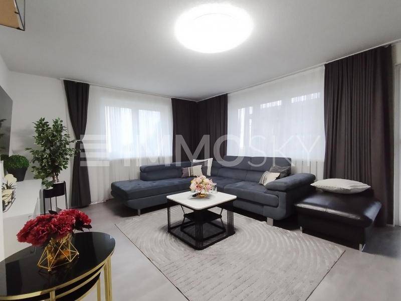 Helles Wohnzimmer mit modernem Sofa, Teppich und dunklen Vorhängen, gut beleuchtet.