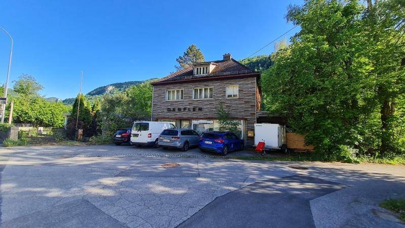 Älteres Gebäude mit traditioneller Fassade und geparkten Autos auf dem Vorplatz vor einer Bergkulisse.