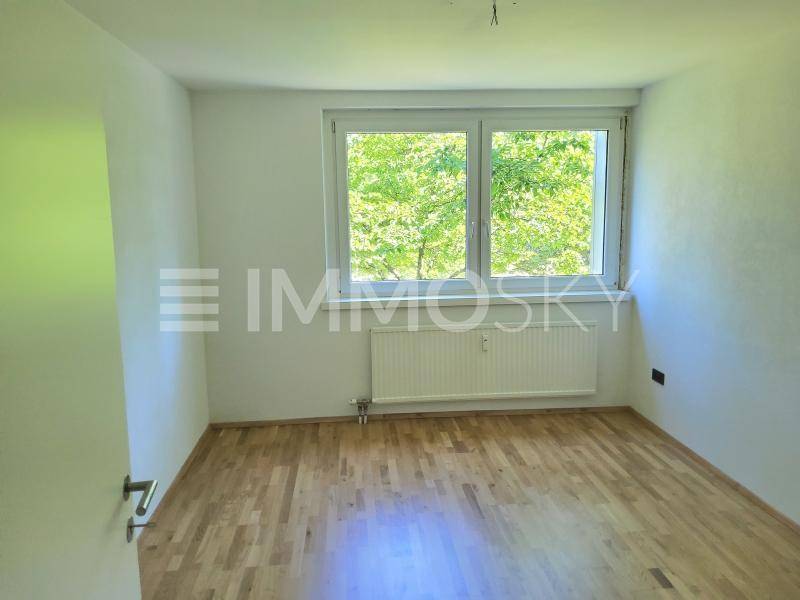 Helles, unmöbliertes Schlafzimmer mit Parkettboden und einem Fenster mit Blick ins Grüne.