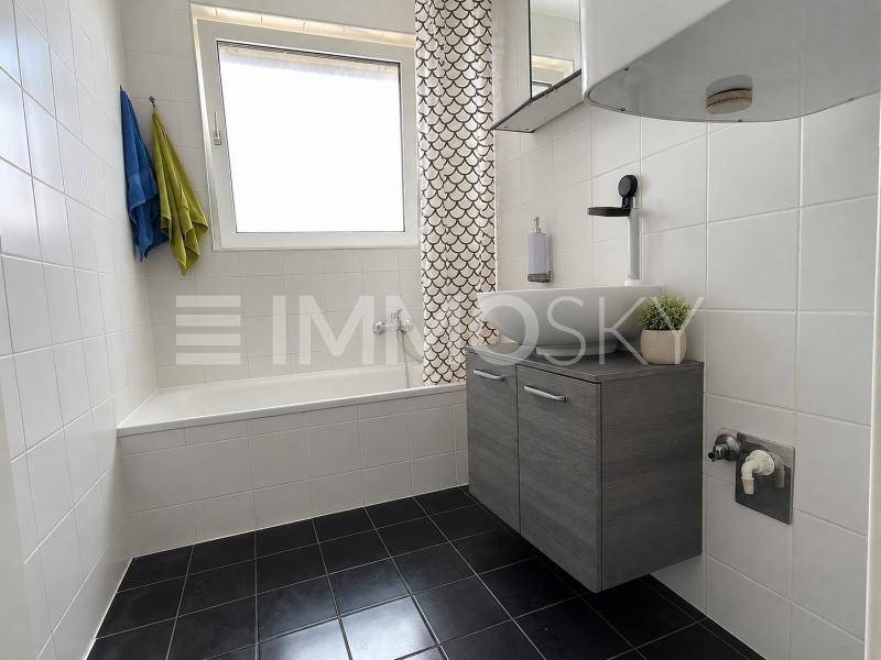 Modernes Badezimmer mit Badewanne, einem einzelnen Waschbecken, Spiegelschrank und einem Fenster für Tageslicht.