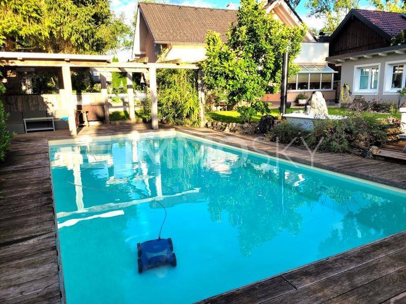 Klarer Swimmingpool mit Poolreiniger und umlaufendem Holzdeck im sonnigen Gartenbereich.