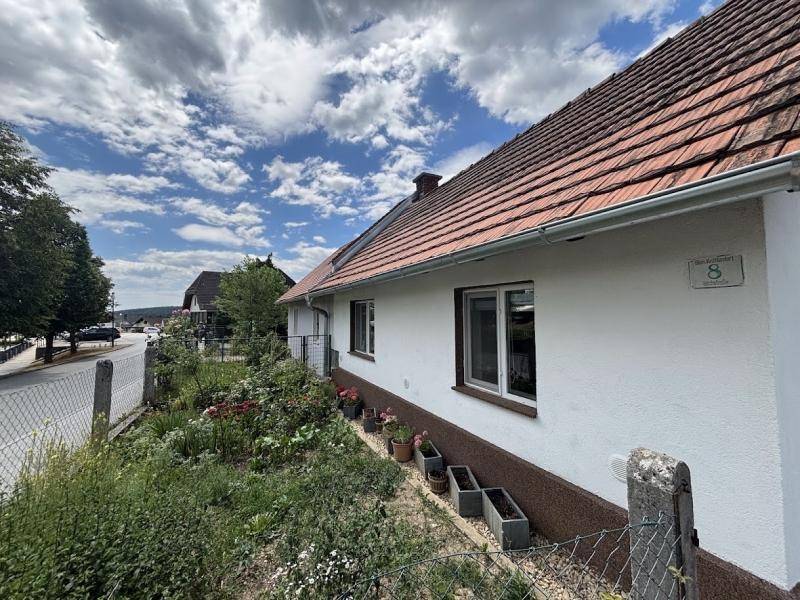 Einfamilienhaus mit weißer Fassade, rotem Ziegeldach und kleinem Vorgarten an einer Straße.
