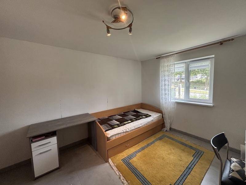 Schlichtes Schlafzimmer mit Einzelbett, Schreibtisch und Fenster mit Vorhängen.