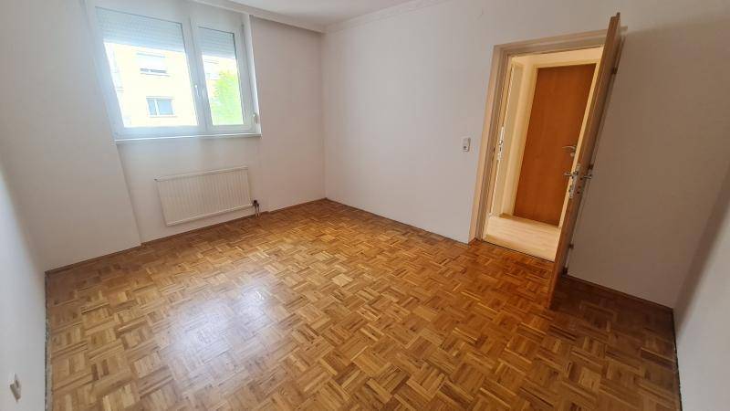 Leeres Schlafzimmer mit Parkettboden und einem Fenster, das viel Tageslicht hereinlässt.