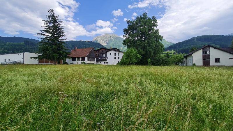 Offene Wiesenfläche mit Blick auf umliegende Häuser und die Berglandschaft im Hintergrund.