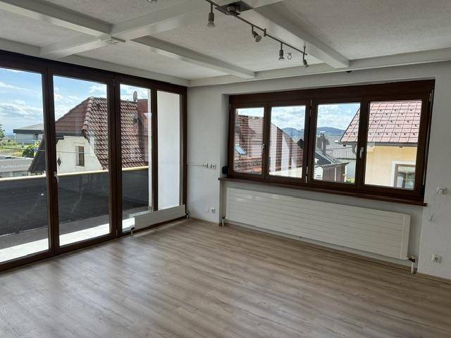 Heller Wohnraum mit großen Fenstern, Zugang zu einem Außenbereich und sichtbaren Deckenbalken.