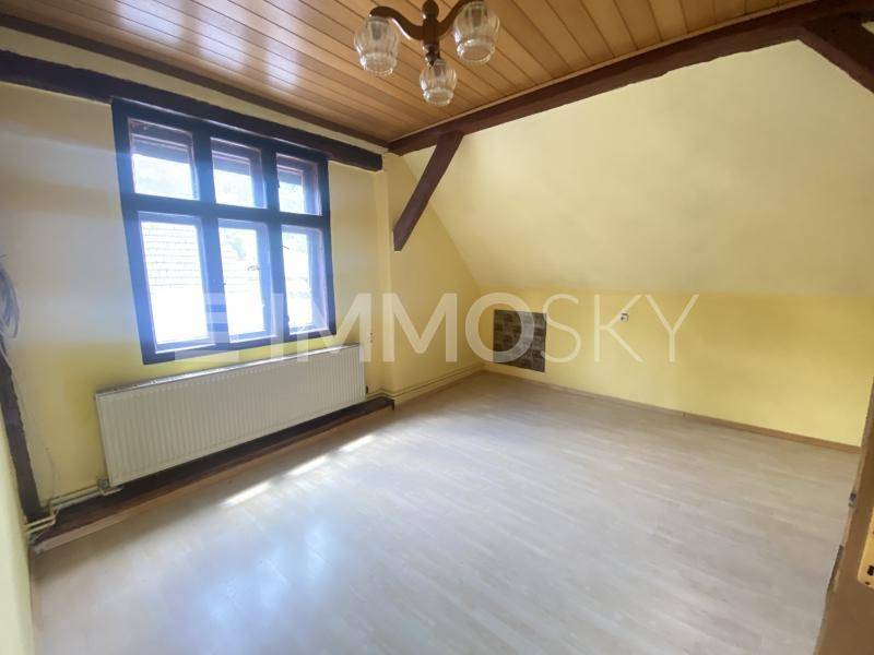 Helles Zimmer mit Holzbalkendecke, Laminatboden und einem Fenster mit Heizkörper darunter.