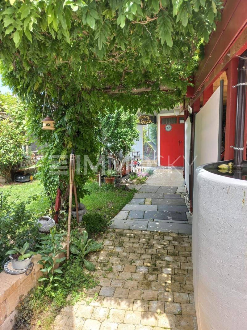 Gepflasterter Gartenweg unter einer grünen Pergola, der zu einem roten Eingang führt.