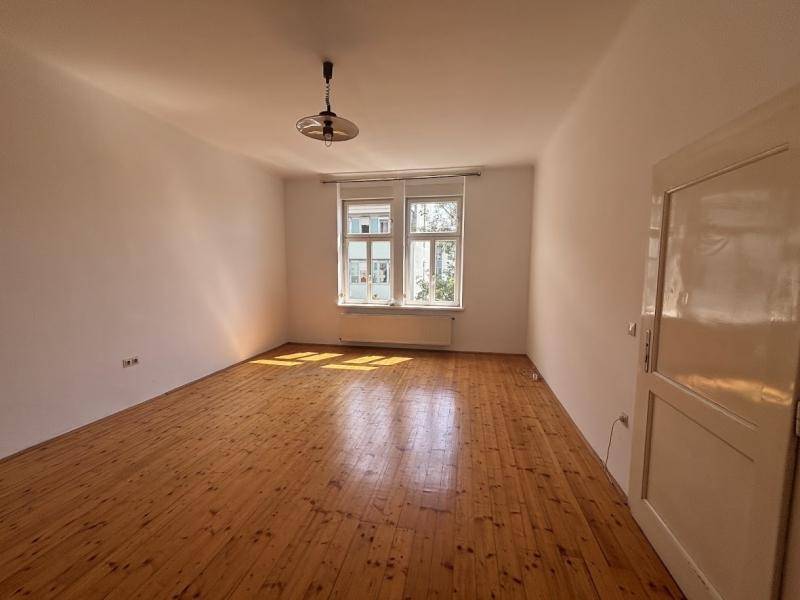 Heller, leerer Raum mit Holzdielenboden, weißen Wänden und einem großen Fenster, das viel Tageslicht hereinlässt.