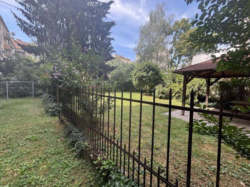 Grüner Gemeinschaftsgarten mit Rasenfläche, hohen Bäumen, Sträuchern und einem kleinen Pavillon im Hintergrund.