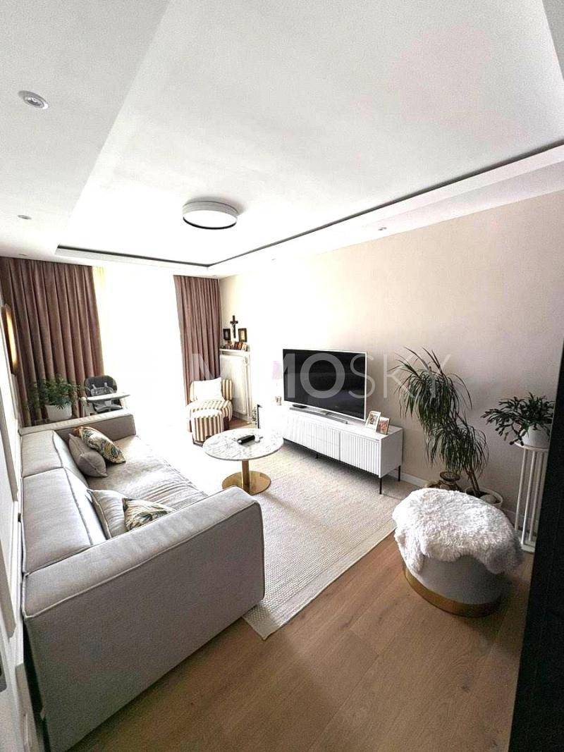 Modernes Wohnzimmer mit beigem Sofa, Fernseher und hellem Holzboden.
