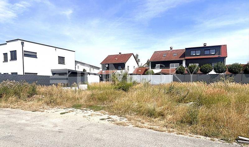 Weitläufiges unbebautes Grundstück mit hohem Gras und verschiedenen Wohnhäusern im Hintergrund.