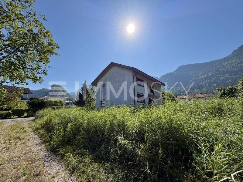 Sonnenbeschienenes Haus mit einfacher Architektur, umgeben von hohem Gras und einer beeindruckenden Bergkulisse.