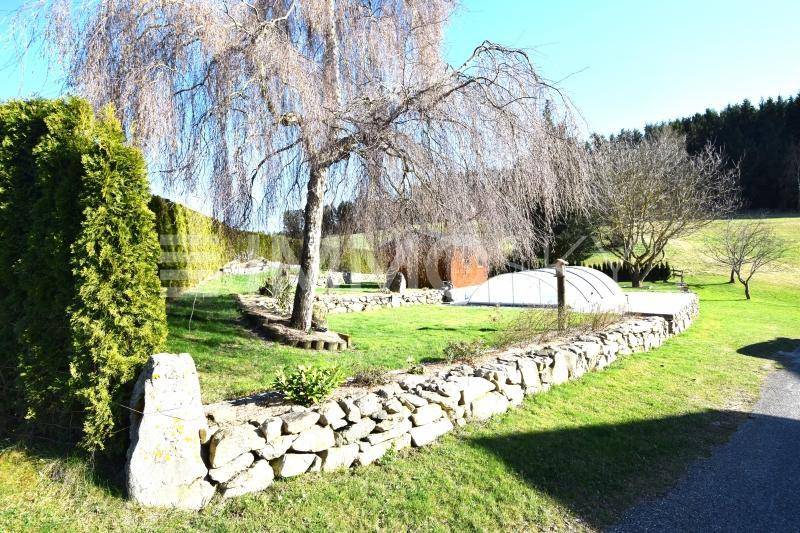 Gepflegter Gartenbereich mit Trauerweide, Steinmauer und einem überdachten Pool im Hintergrund.