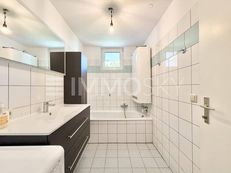 Gefliestes Badezimmer mit Badewanne, Einzelwaschbecken und Fenster, ausgestattet mit einem Warmwasserboiler.