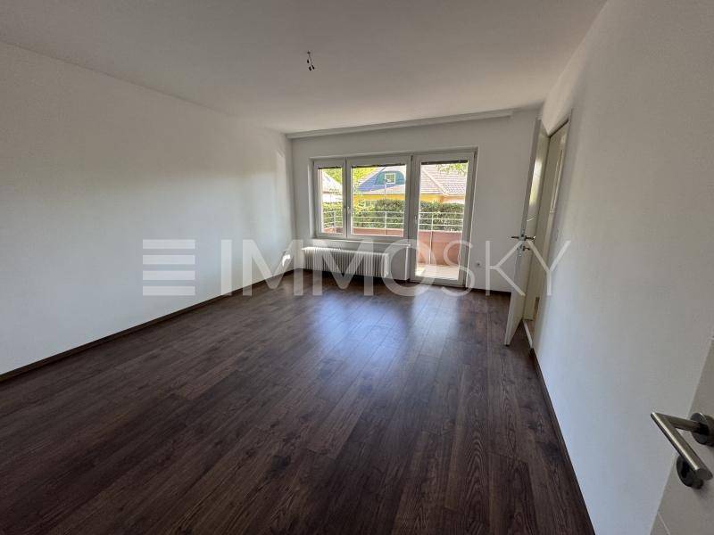 Helles Zimmer mit dunklem Laminatboden und Blick ins Grüne durch die Fenster.