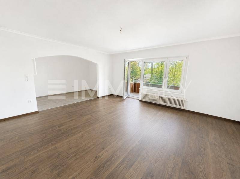 Geräumiges Wohnzimmer mit dunklem Laminatboden, großen Fenstern und Zugang zum Balkon.