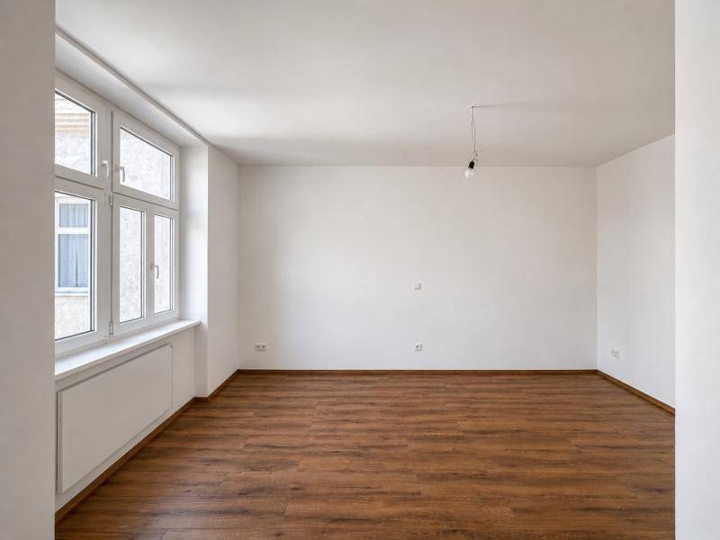 Geräumiges, helles Zimmer mit Holzboden und großem Fenster, das viel Tageslicht hereinlässt.