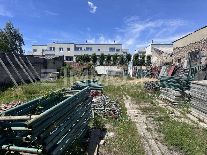 Überwachsenes Baugrundstück mit Baumaterialien und modernen Gebäuden im Hintergrund.