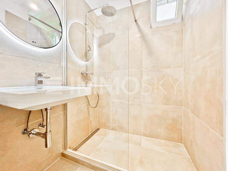 Modernes Badezimmer mit beigefarbenen Fliesen, begehbarer Dusche und beleuchteten Spiegeln.