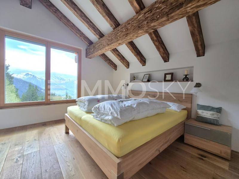 Schlafzimmer mit Holzbalken, großem Fenster und Panoramablick auf die Berge und den See.