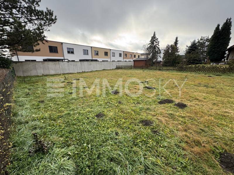 Freies Baugrundstück mit Gras und Bäumen, einer Betonmauer und Reihenhäusern im Hintergrund unter bewölktem Himmel.