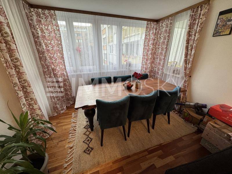 Gemütlicher Essbereich mit Holztisch, grünen Samtstühlen und großen Fenstern mit gemusterten Vorhängen.