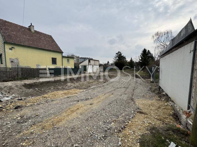 Schotterweg auf einem unbebauten Grundstück, flankiert von älteren Häusern unter trübem Himmel.
