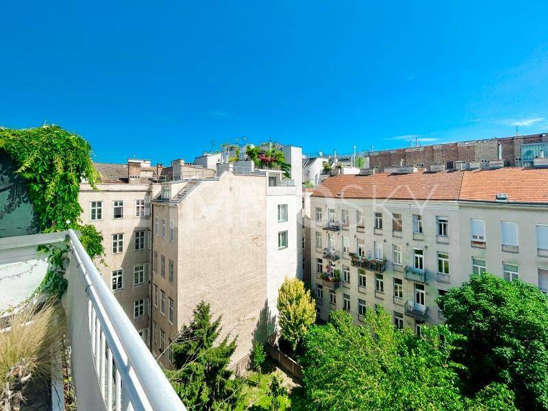 Blick von einem Balkon auf umliegende Gebäude und grüne Bäume unter blauem Himmel.