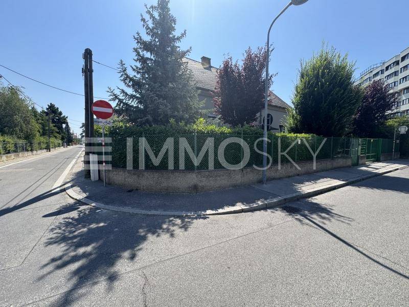 Eckansicht des Grundstücks von der Straße aus mit Haus, Zaun und umgebender Vegetation.