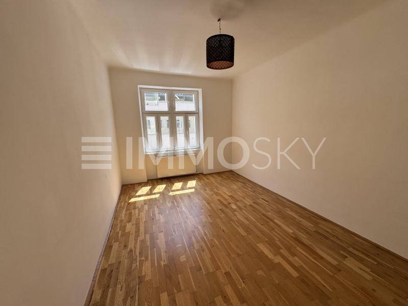 Helles Schlafzimmer mit Holzboden, weißen Wänden und einem Fenster, durch das Sonnenlicht einfällt.