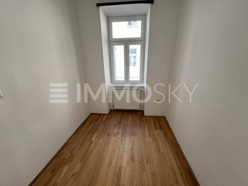 Leerer Raum mit Holzboden, weißen Wänden und einem hohen, schmalen Fenster mit Heizkörper.