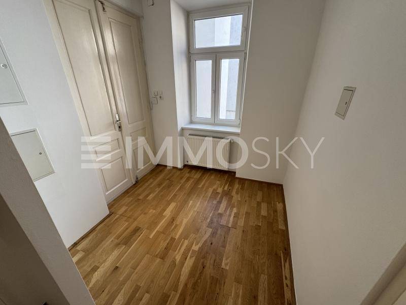 Leerer Raum mit Holzboden, weißen Wänden und einem Fenster mit Heizkörper darunter.