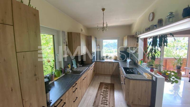 Helle Küche mit Holzschränken, dunklen Arbeitsplatten und Blick ins Grüne durch große Fenster.
