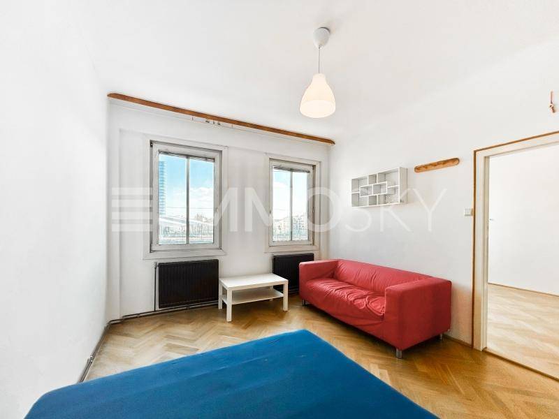 Wohnzimmer mit rotem Sofa, Fischgrätparkett und zwei Fenstern mit Stadtblick.