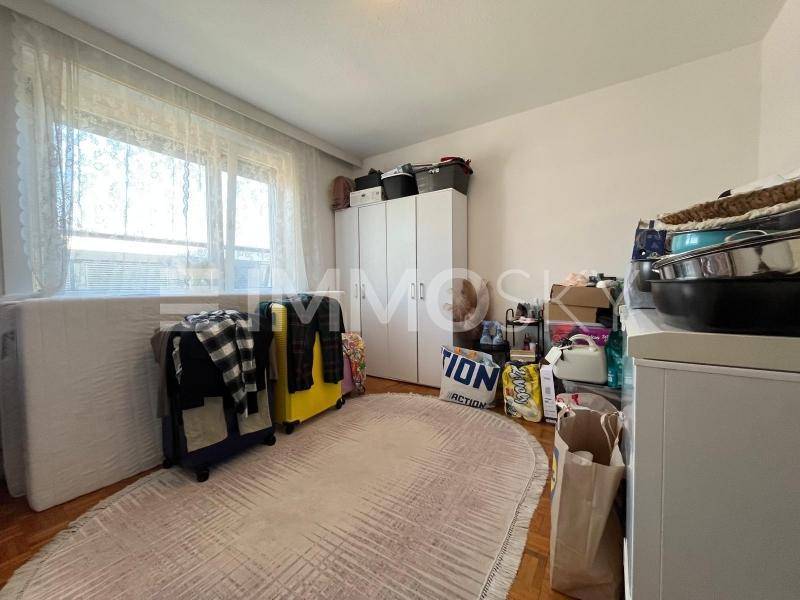 Helles Schlafzimmer mit großem Fenster, Teppich und verschiedenen Aufbewahrungsmöbeln.