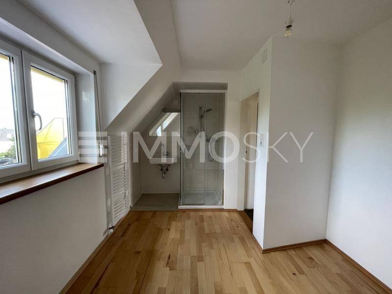 Heller Raum mit Holzboden, Dachschräge und integrierter Duschkabine neben dem Fenster.