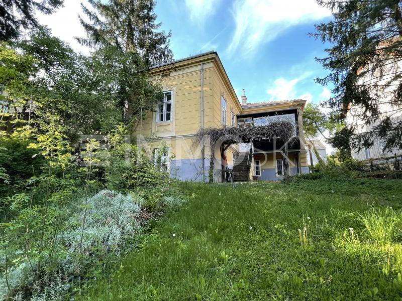 Historisches gelbes Gebäude mit überdachtem Balkon, umgeben von üppiger grüner Vegetation und Bäumen.