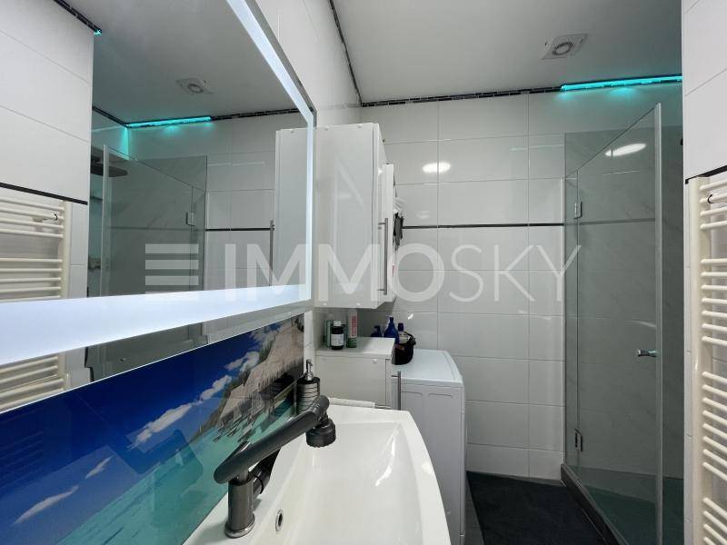Modernes Badezimmer mit Dusche, Einzelwaschbecken und Waschmaschine, akzentuiert durch blaue Beleuchtung.