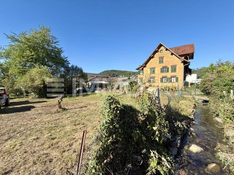 Traditionelles Haus mit gelber Fassade und grünen Fensterläden, umgeben von einem sonnigen Grundstück mit Bach.