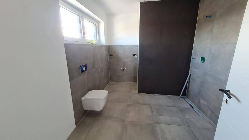 Modernes Badezimmer im Rohbau mit grauen Fliesen, wandhängendem WC und Fenster.