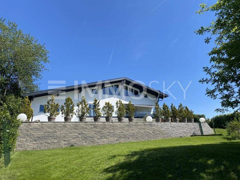 Moderne weiße Villa mit großen Fenstern, Balkon und gepflegtem Gartenbereich unter blauem Himmel.